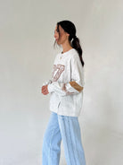 A woman wearing a light gray marled sweatshirt with a brown patterned graphic and light blue wide-leg jeans, standing against a plain off-white wall.