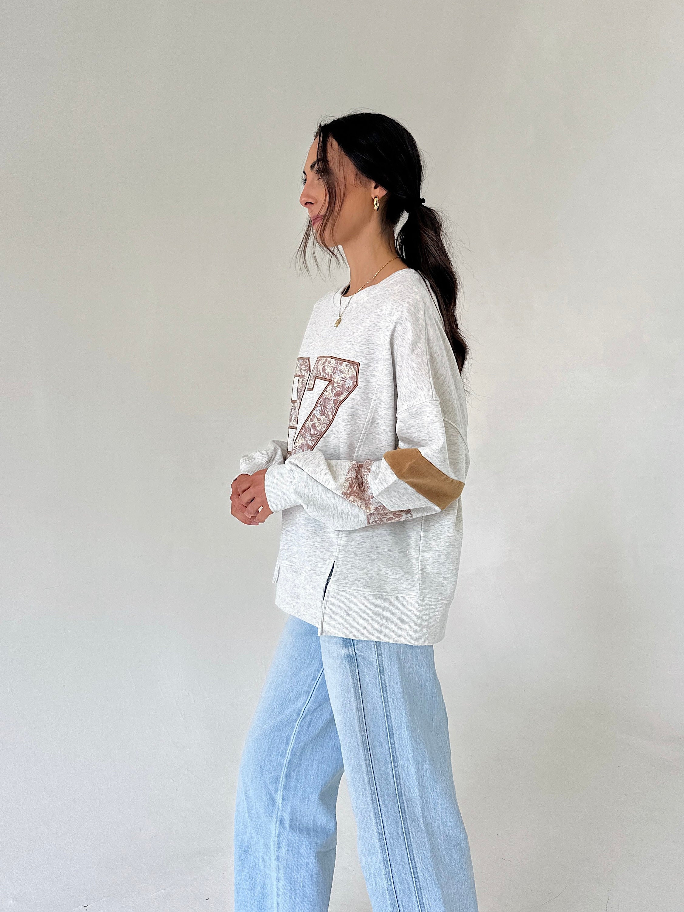 A woman wearing a light gray marled sweatshirt with a brown patterned graphic and light blue wide-leg jeans, standing against a plain off-white wall.