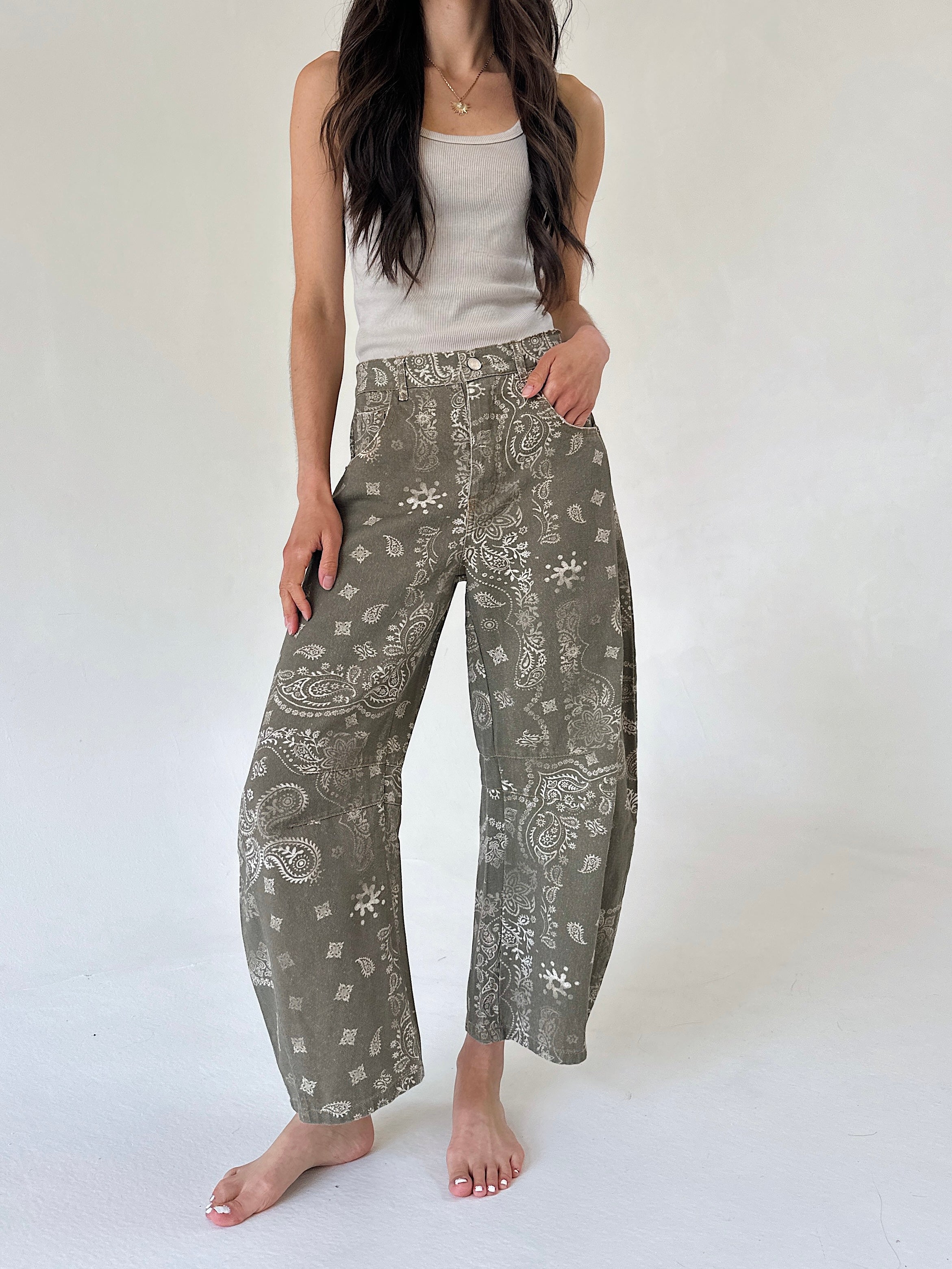 A woman modeling wide-leg barrel pants in an olive green paisley and floral print, paired with a white ribbed tank top, standing against a plain white studio background.