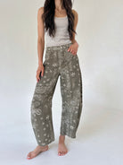 A woman modeling wide-leg barrel pants in an olive green paisley and floral print, paired with a white ribbed tank top, standing against a plain white studio background.