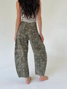 A person shown from the back wearing loose, wide-leg pants with an olive green and white paisley pattern, paired with a light gray sleeveless top, standing barefoot against a plain white studio background.