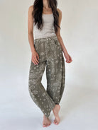 A woman modeling a pair of loose, wide-leg pants in an olive green paisley pattern, paired with a light beige tank top, against a plain off-white studio background.
