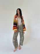 A woman stands barefoot against a plain light gray studio background, wearing a multicolor striped crochet cardigan with wide sleeves over a beige tank top and olive green patterned wide-leg barrel pants.