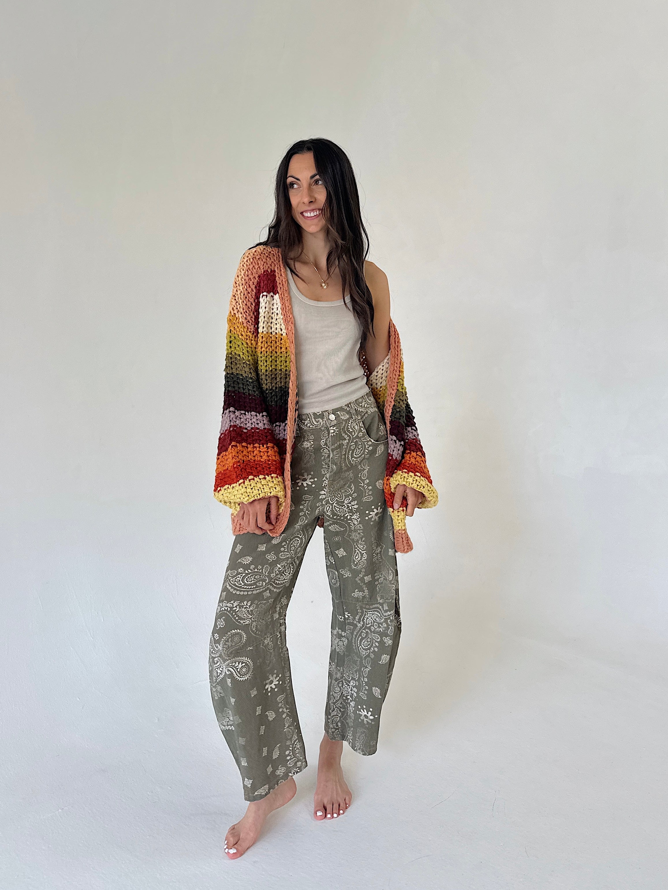 A woman stands barefoot against a plain light gray studio background, wearing a multicolor striped crochet cardigan with wide sleeves over a beige tank top and olive green patterned wide-leg barrel pants.