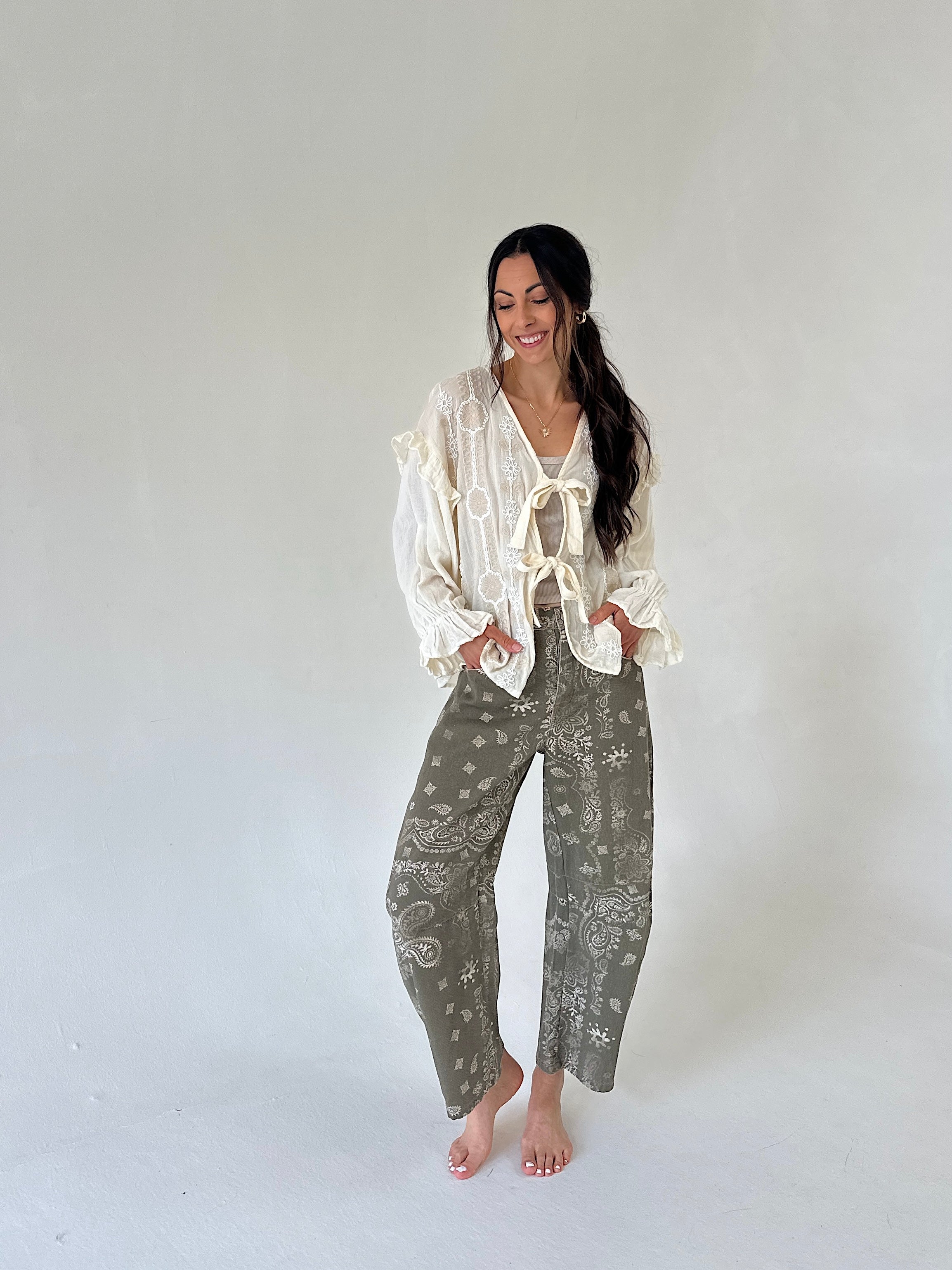 A woman modeling a bohemian-style outfit consisting of a cream lace blouse with ruffles and tie details, paired with olive green paisley bandana-print pants, standing against a plain light gray studio background.