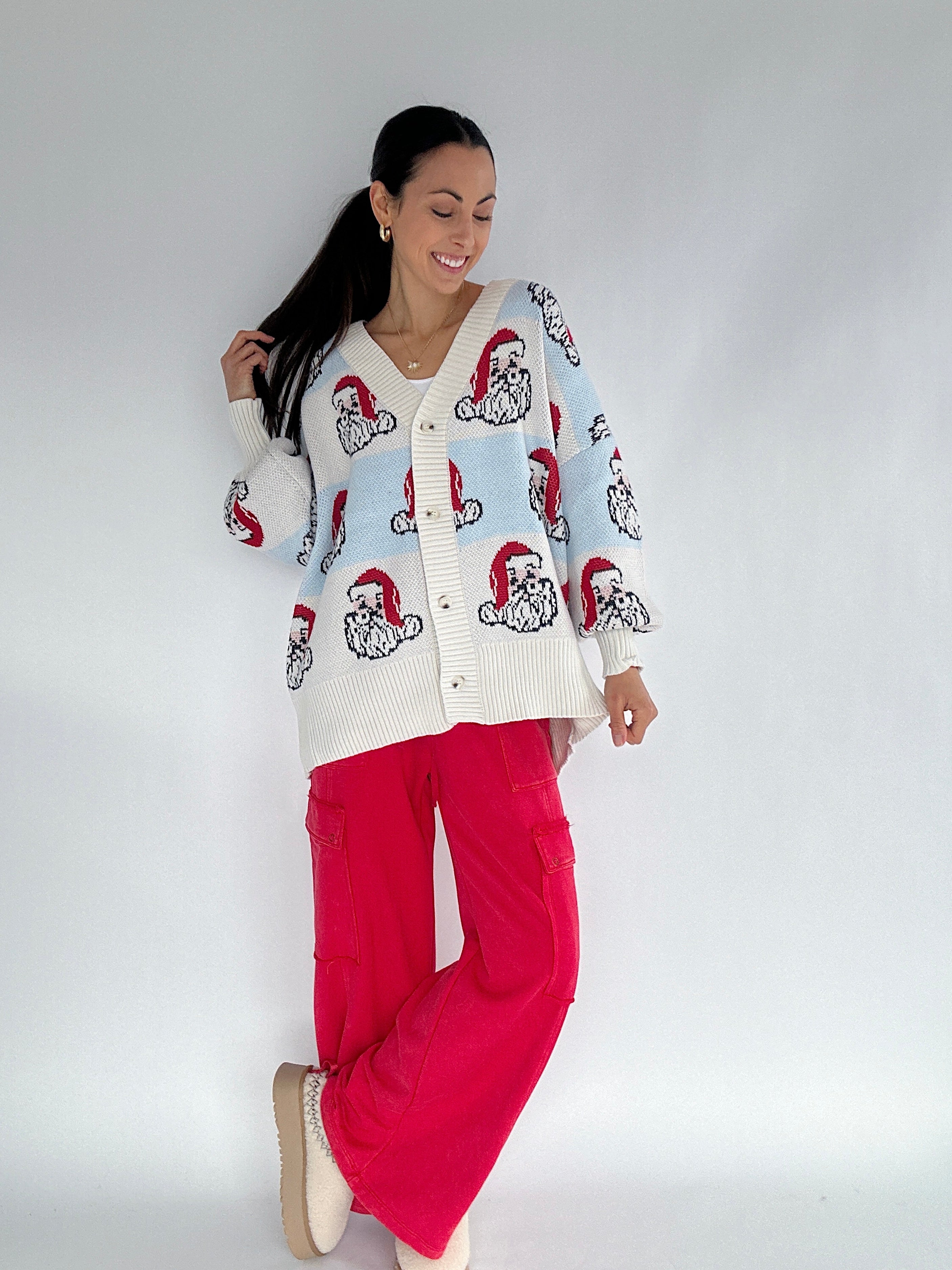 Woman wearing a white cardigan with red and blue stripe and Santa face pattern and red pants on a white background