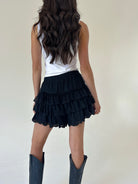 A woman shown from the back wearing black ruffled tiered shorts, a white sleeveless top, and dark cowboy boots against a plain off-white studio background.