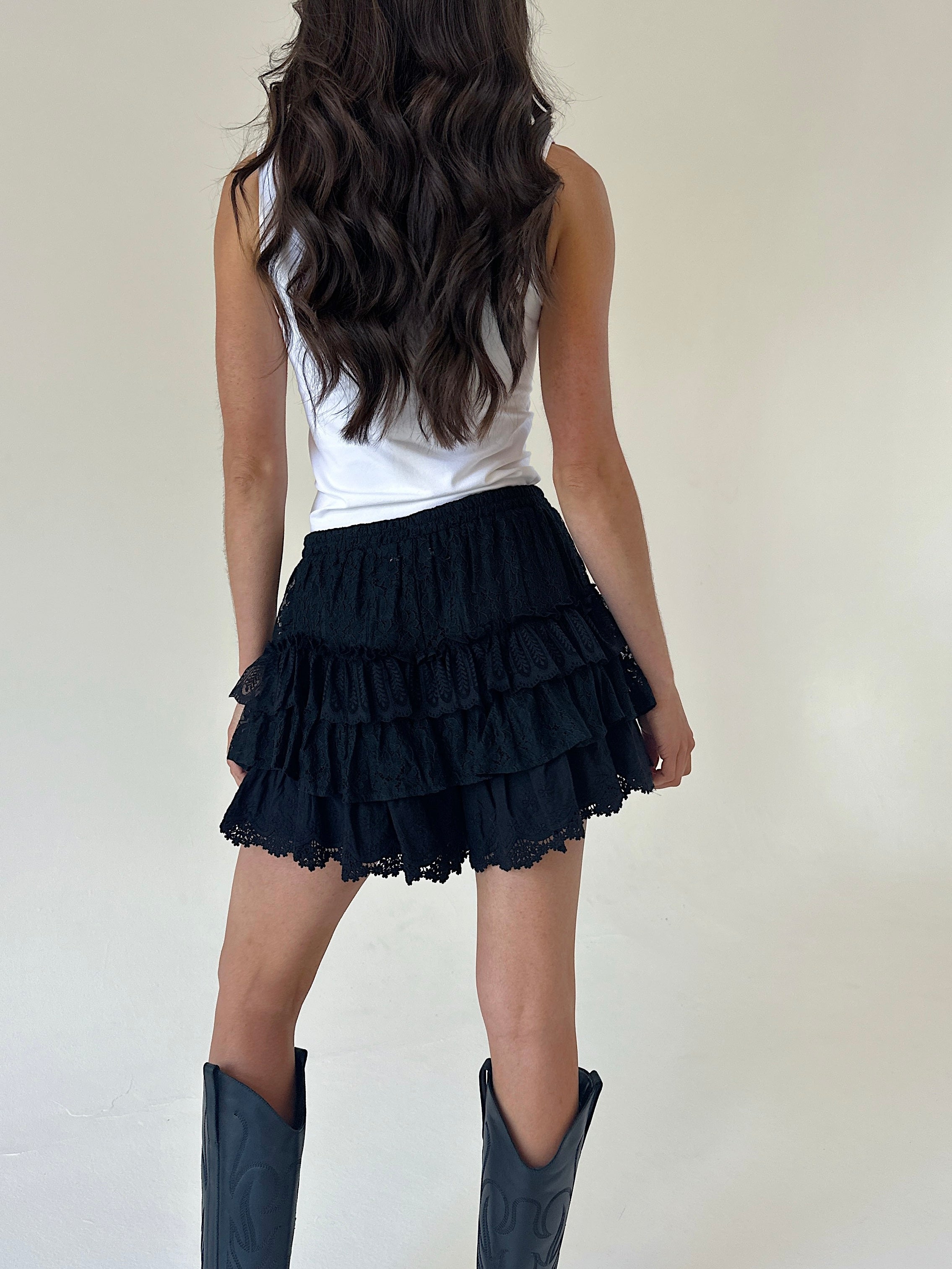 A woman shown from the back wearing black ruffled tiered shorts, a white sleeveless top, and dark cowboy boots against a plain off-white studio background.