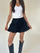 A woman wearing black ruffled tiered shorts with lace trim, paired with a white tank top and black cowboy boots, standing against a plain off-white wall.