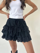 A model wearing black tiered ruffle shorts with a lace hem and drawstring, paired with a white tank top.