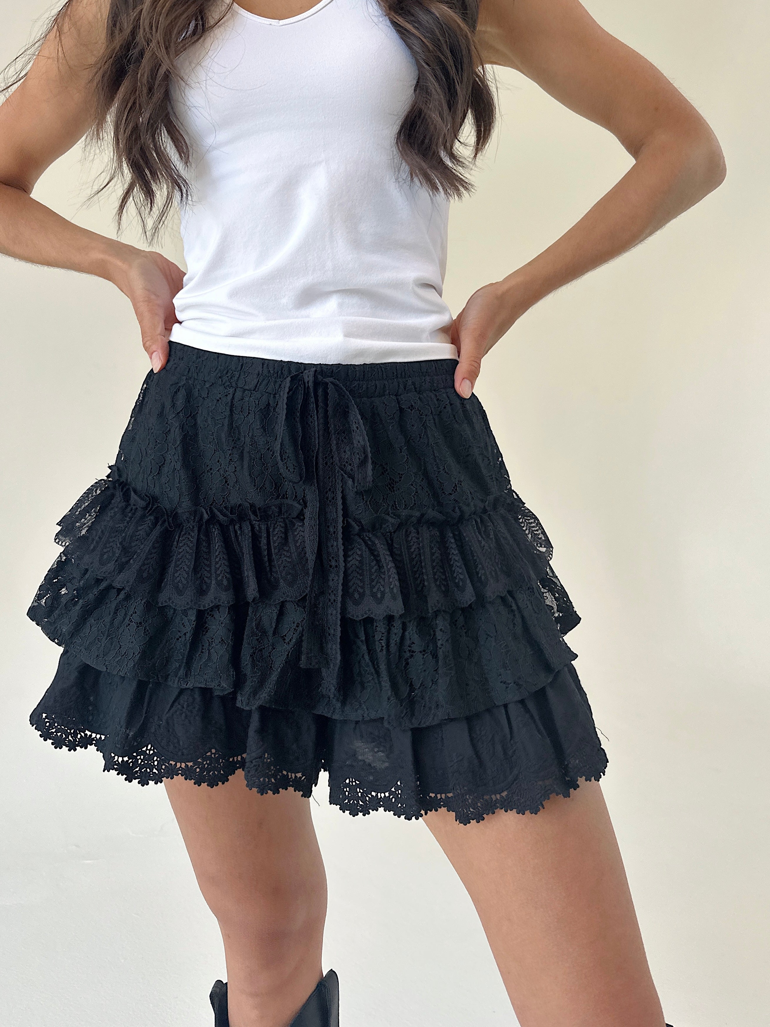 A model wearing black tiered ruffle shorts with a lace hem and drawstring, paired with a white tank top.