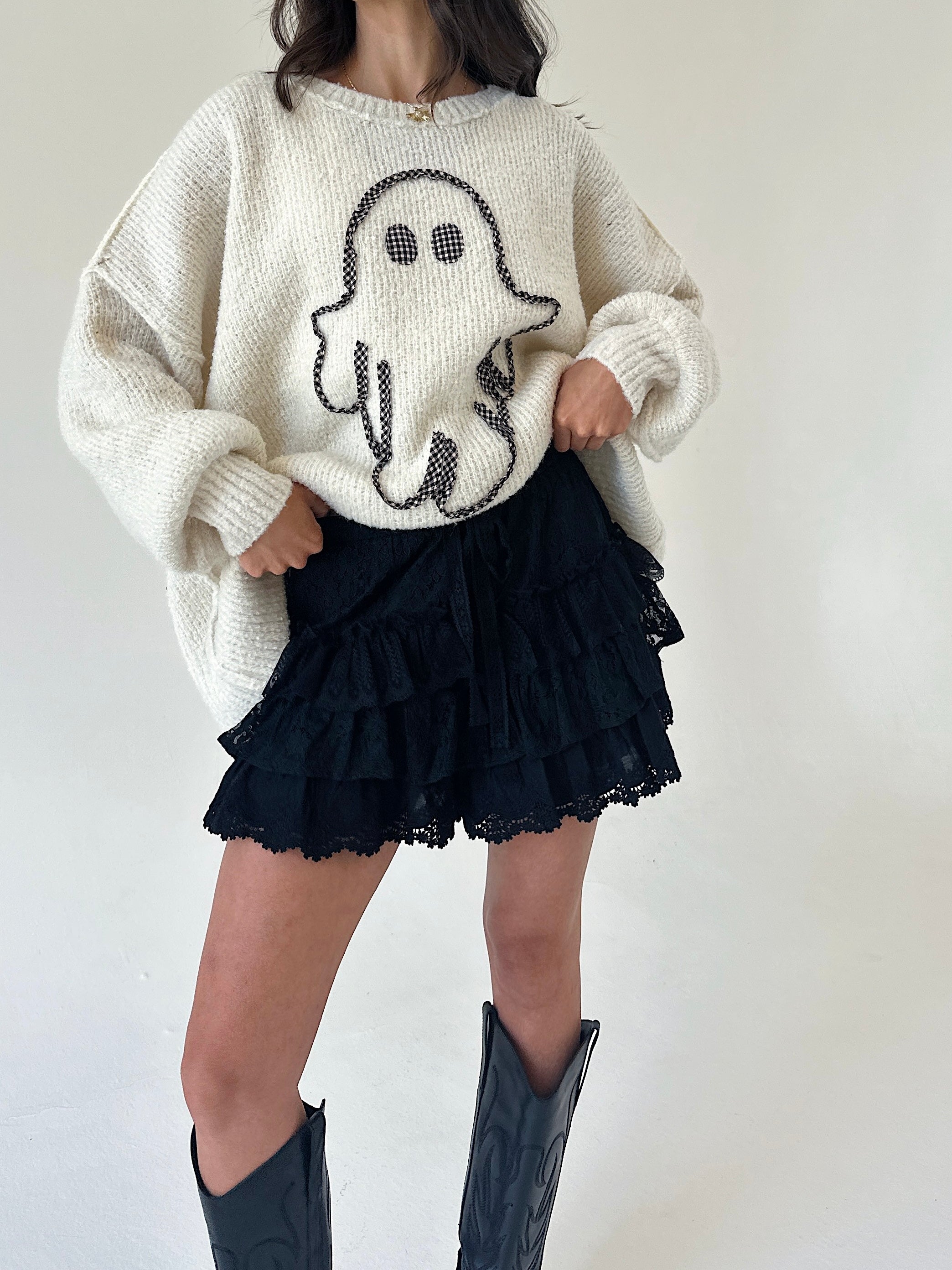 A person wearing a cream knit sweater with a ghost motif, black ruffled shorts, and black cowboy boots, standing against a plain light background.