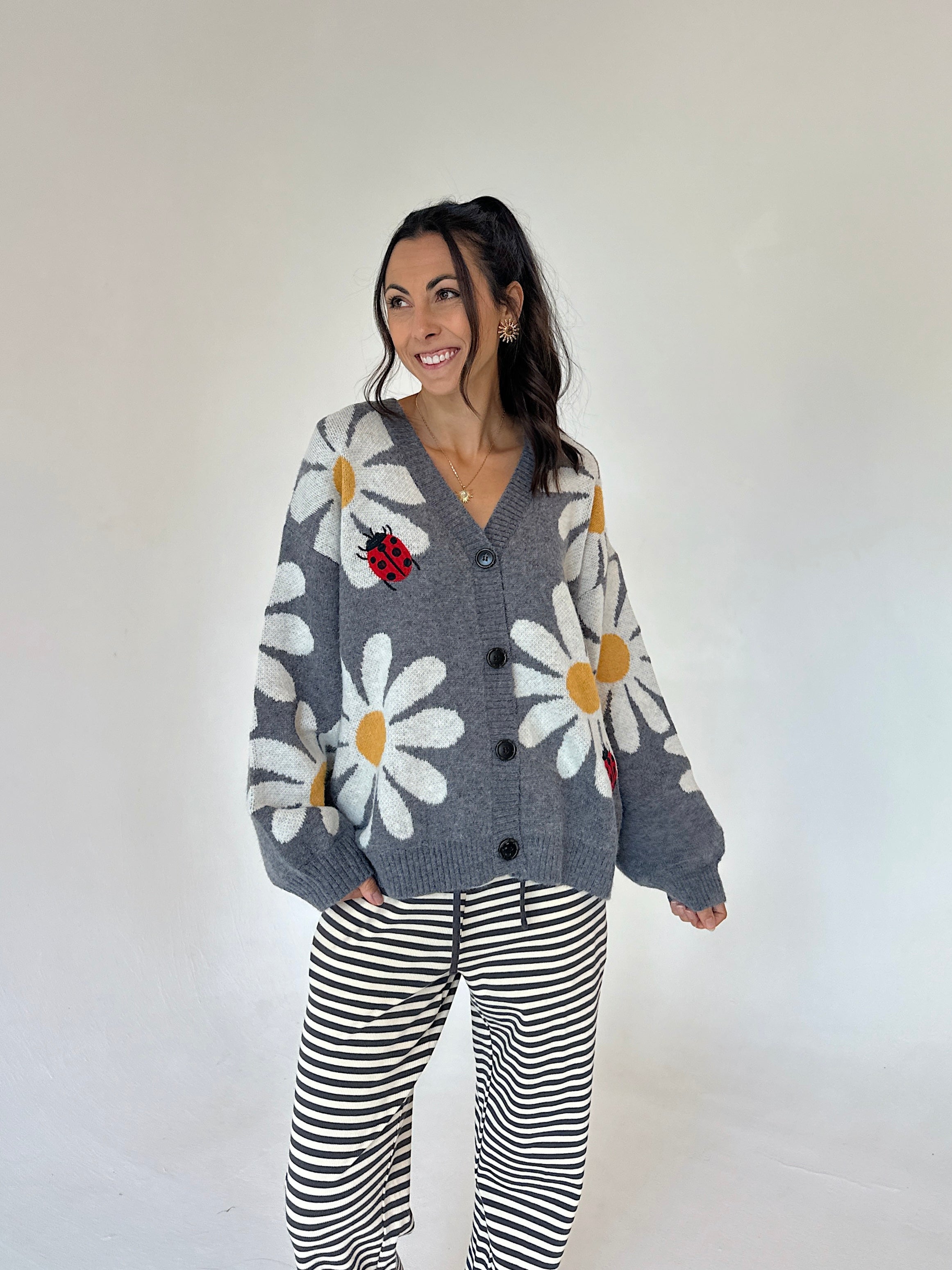 A woman modeling a gray knit cardigan with large white daisy patterns, yellow centers, and a red ladybug applique, paired with black and white striped pants, against a plain off-white studio background.