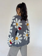 A woman viewed from behind wearing a gray knit sweater with large white daisy patterns, yellow centers, and red ladybugs, paired with black and white striped pants against a plain off-white background.