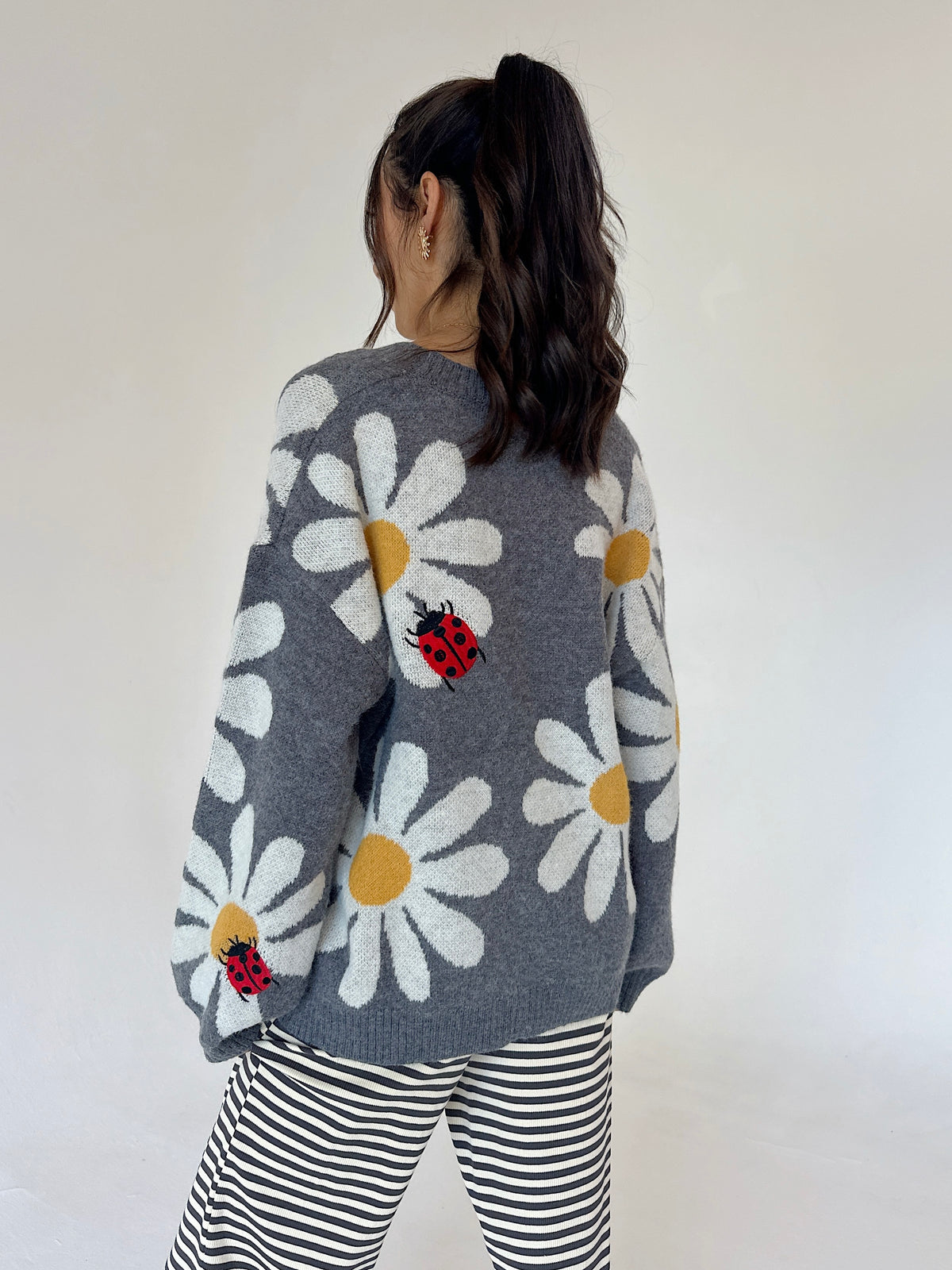 A woman viewed from behind wearing a gray knit sweater with large white daisy patterns, yellow centers, and red ladybugs, paired with black and white striped pants against a plain off-white background.