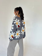A woman is shown from the back wearing a gray knit sweater with large white daisy patterns, yellow centers, and small red ladybug appliqués, paired with black and white striped pants, against a plain off-white background.