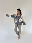 A woman modeling a gray knit cardigan and matching striped pants. The cardigan features large white daisy patterns with yellow centers and small red ladybug appliqués.