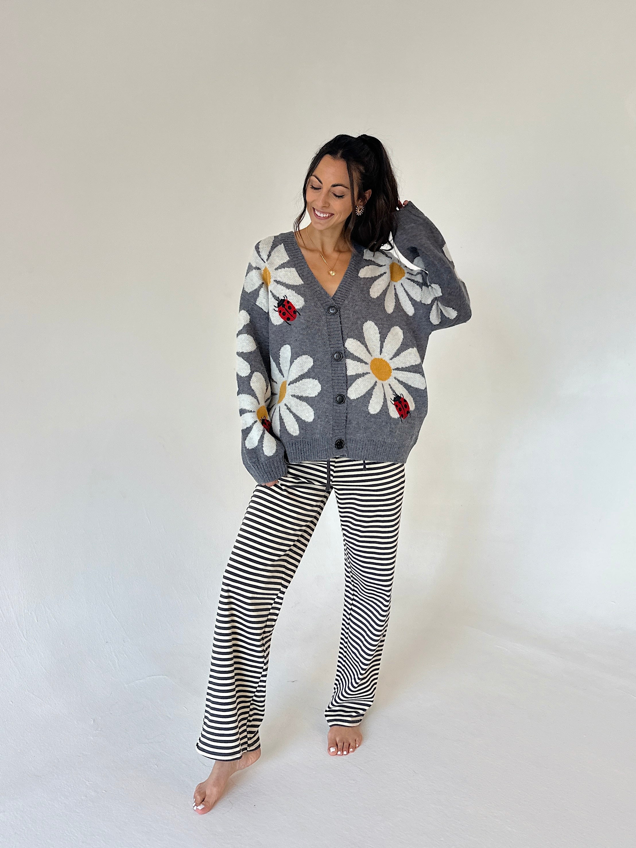 A woman modeling a cozy gray knit cardigan with large white daisy and ladybug motifs, paired with black and white horizontally striped pants, against a plain off-white studio background.