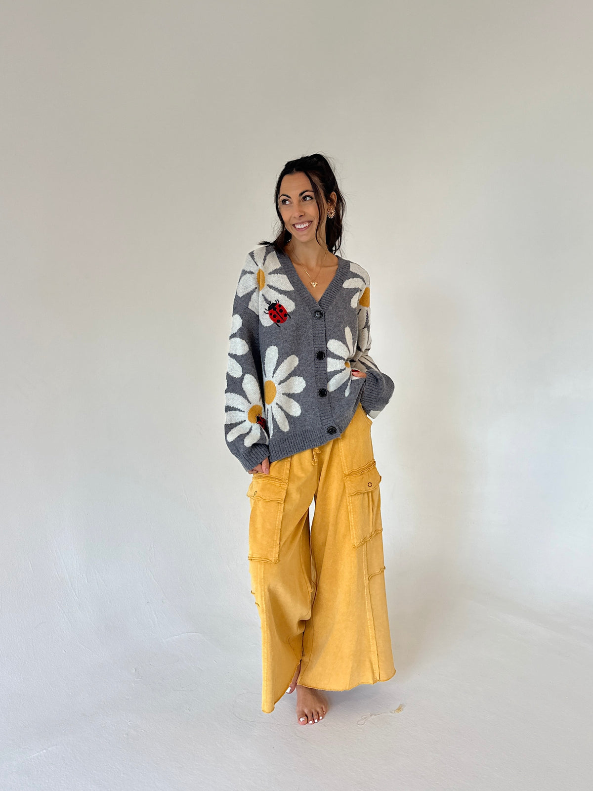 A woman stands barefoot against a plain light gray studio background, wearing a gray knit cardigan with large white daisy patterns and a small red flower accent, paired with wide-leg mustard yellow pants.