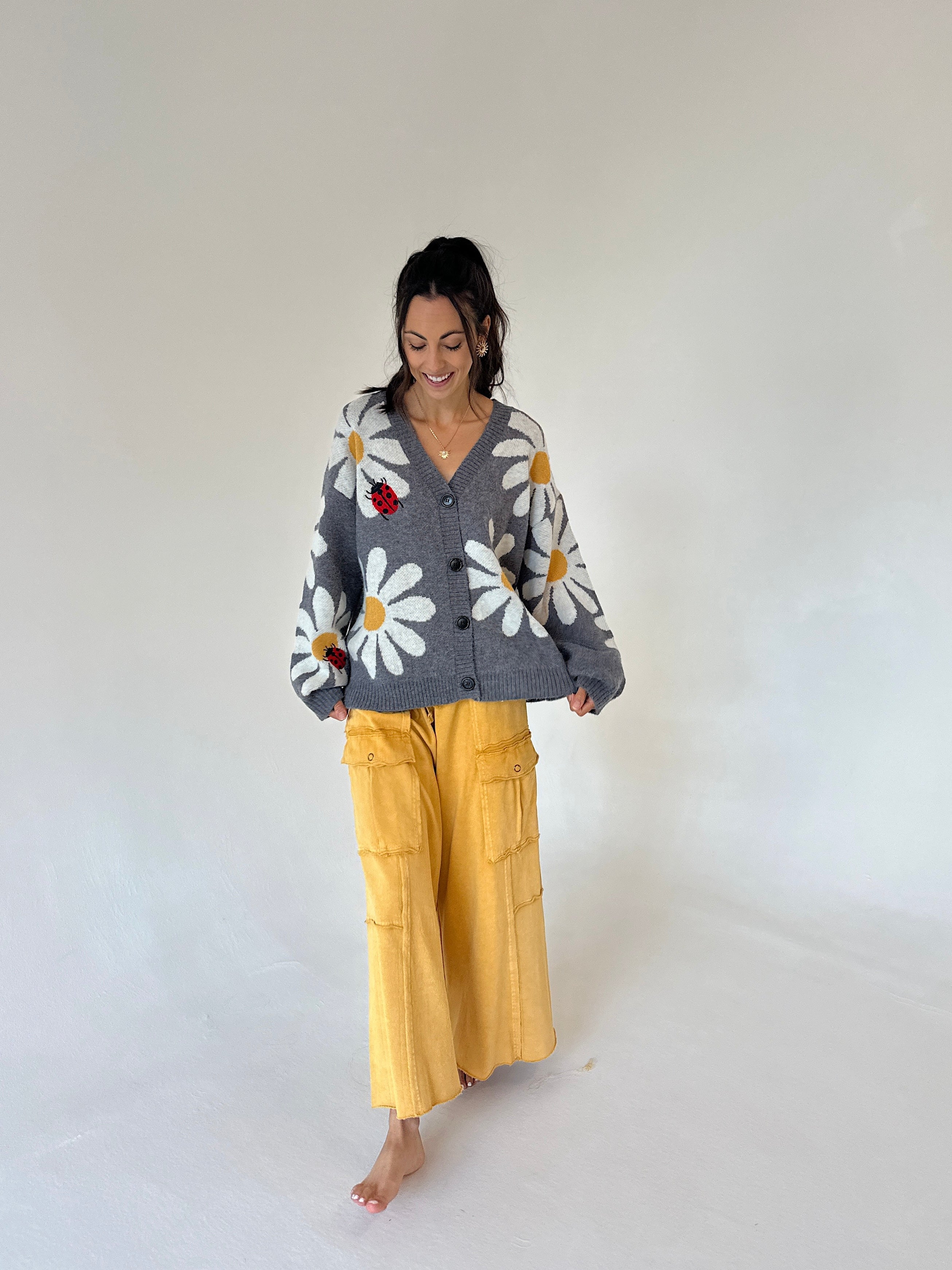 A woman stands barefoot against a plain white studio background, wearing a gray knit cardigan with large white daisy flowers and yellow cargo pants.
