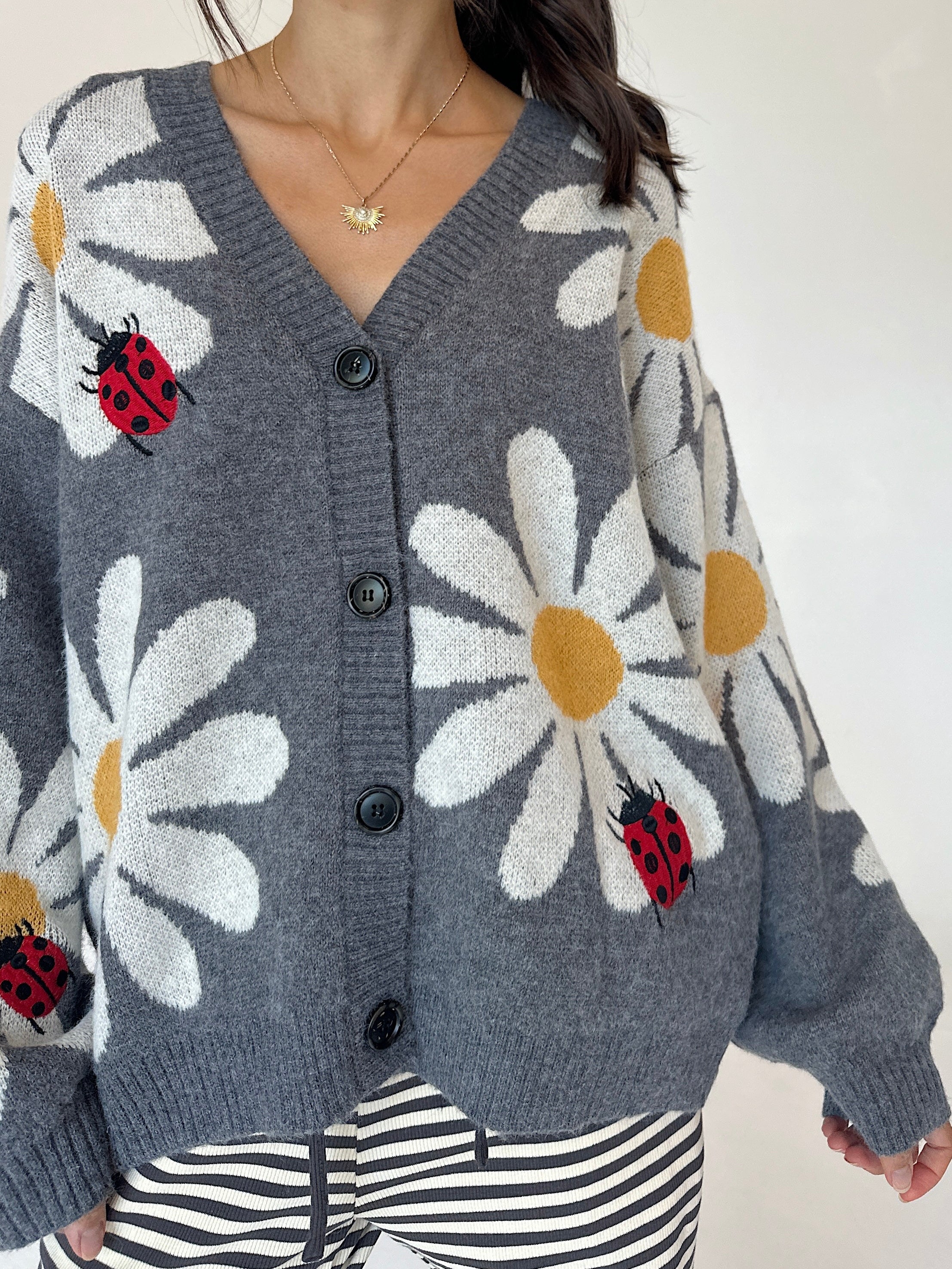 A woman wearing a gray knit cardigan with large white daisy patterns, yellow centers, and red ladybugs, paired with black-and-white striped pants.