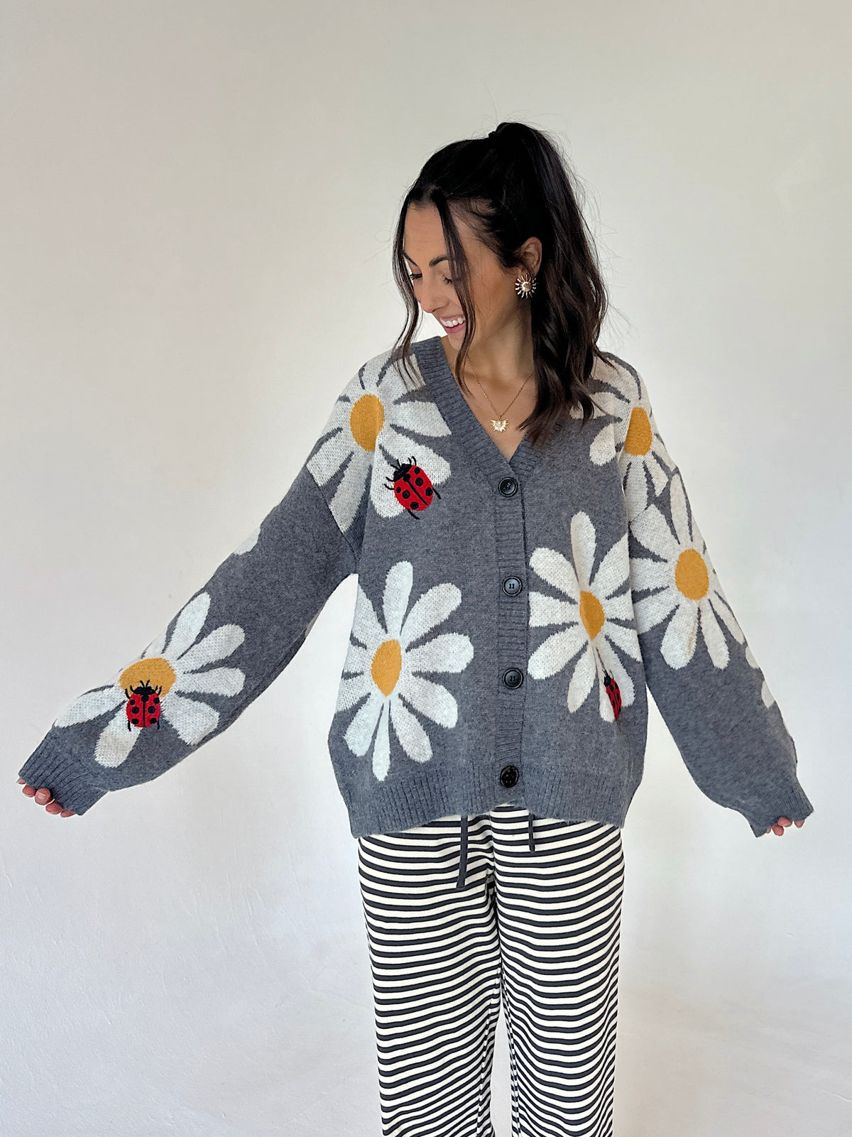 A woman modeling a gray knit cardigan with large white daisy patterns, yellow centers, and red ladybug motifs, paired with black and white striped pants against a plain light background.