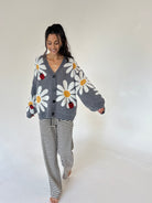 A woman modeling a cozy gray knit cardigan with large white daisy patterns, yellow centers, and small red ladybugs, paired with black and white striped pajama pants, against a plain white studio background.