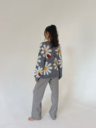 A woman standing with their back to the camera, wearing a gray knit sweater with a daisy and ladybug pattern, paired with black and white horizontally striped pants, against a plain off-white background.