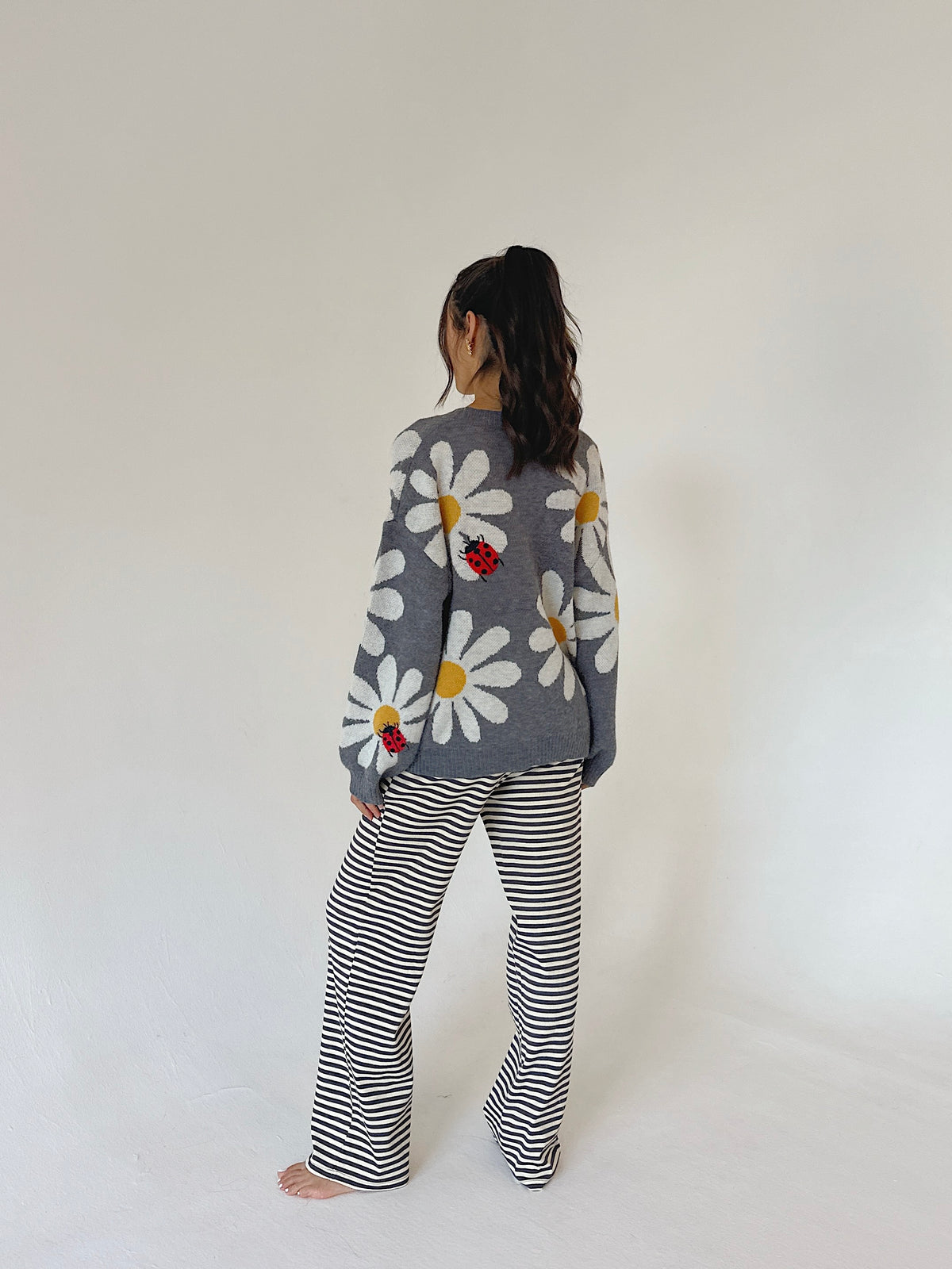 A woman standing with their back to the camera, wearing a gray knit sweater with a daisy and ladybug pattern, paired with black and white horizontally striped pants, against a plain off-white background.