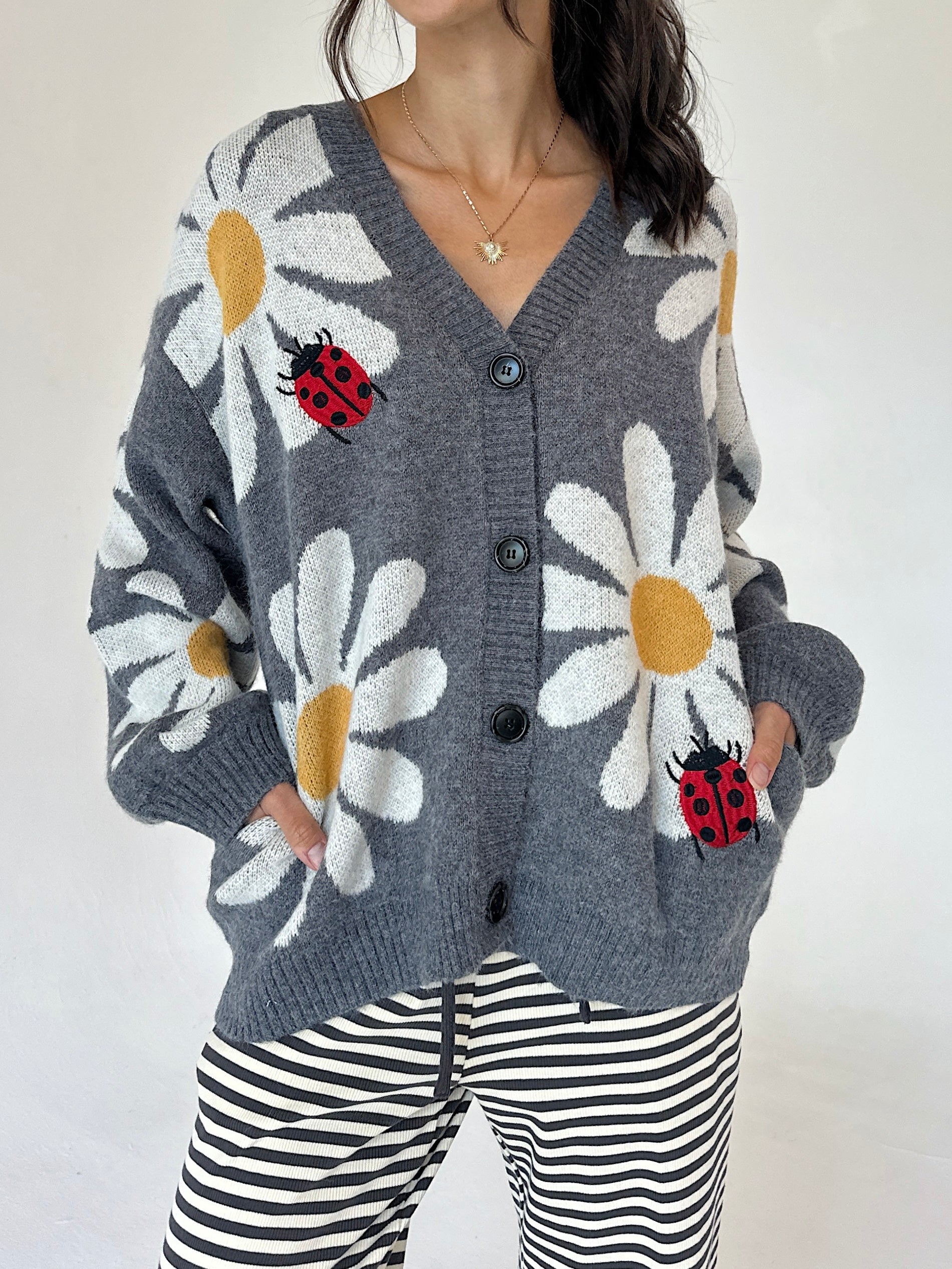 A woman wearing a gray knit cardigan with large white daisy patterns, yellow centers, and red ladybugs, paired with black and white striped pants against a plain light background.