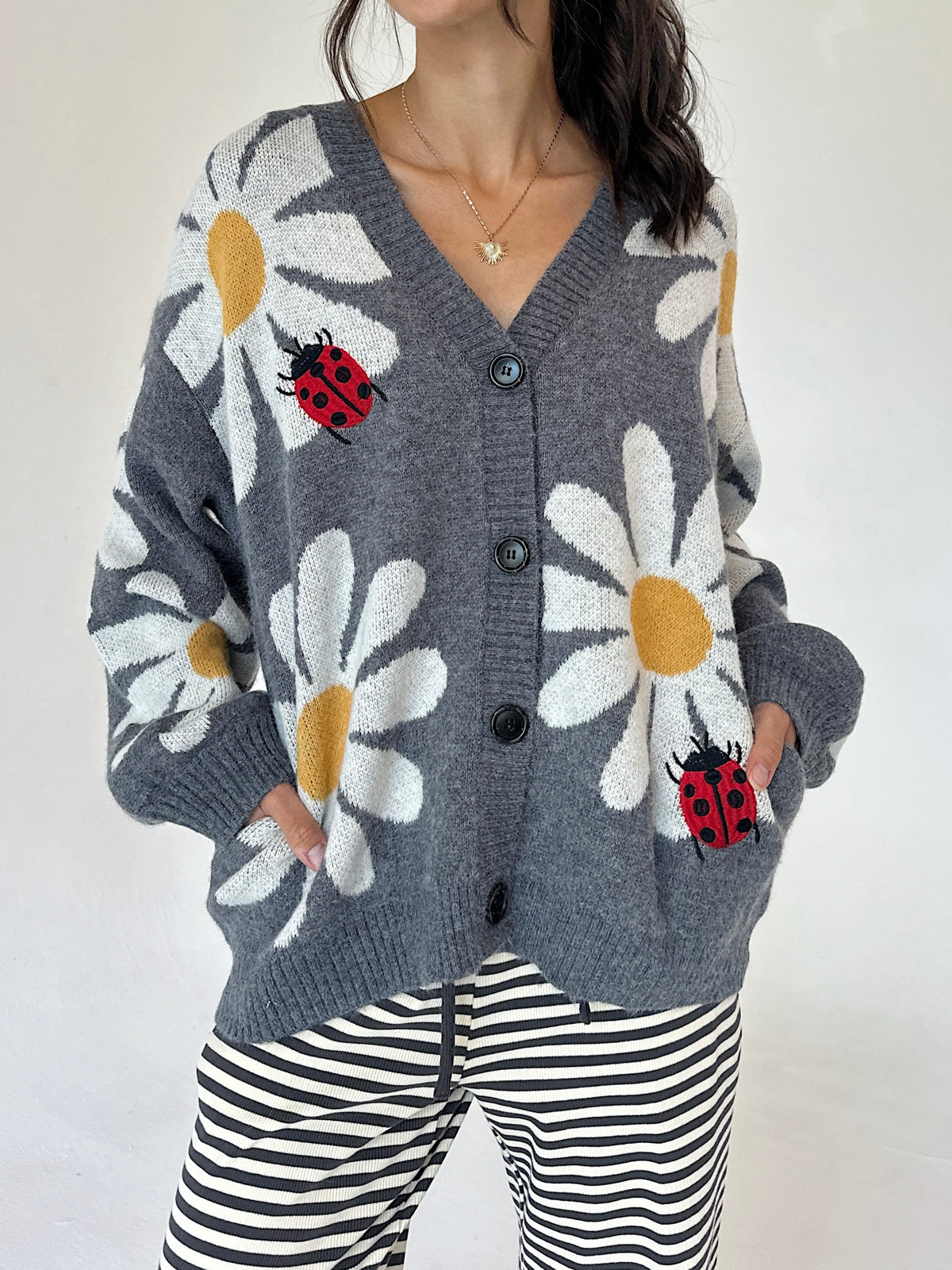 A woman wearing a gray knit cardigan with large white daisy patterns, yellow centers, and red ladybugs, paired with black and white striped pants against a plain light background.