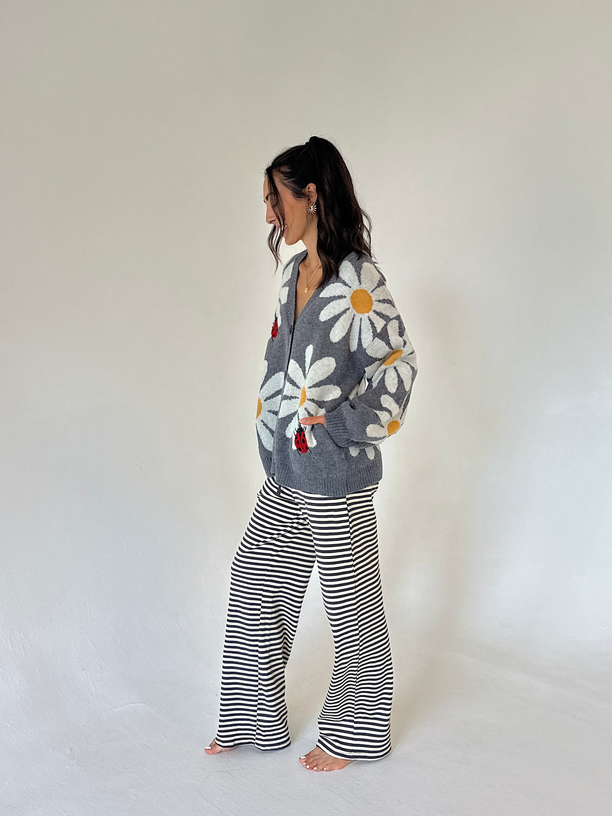 A woman stands barefoot in profile wearing a gray cardigan with a large white daisy print and black-and-white horizontally striped wide-leg pants against a plain off-white studio background.
