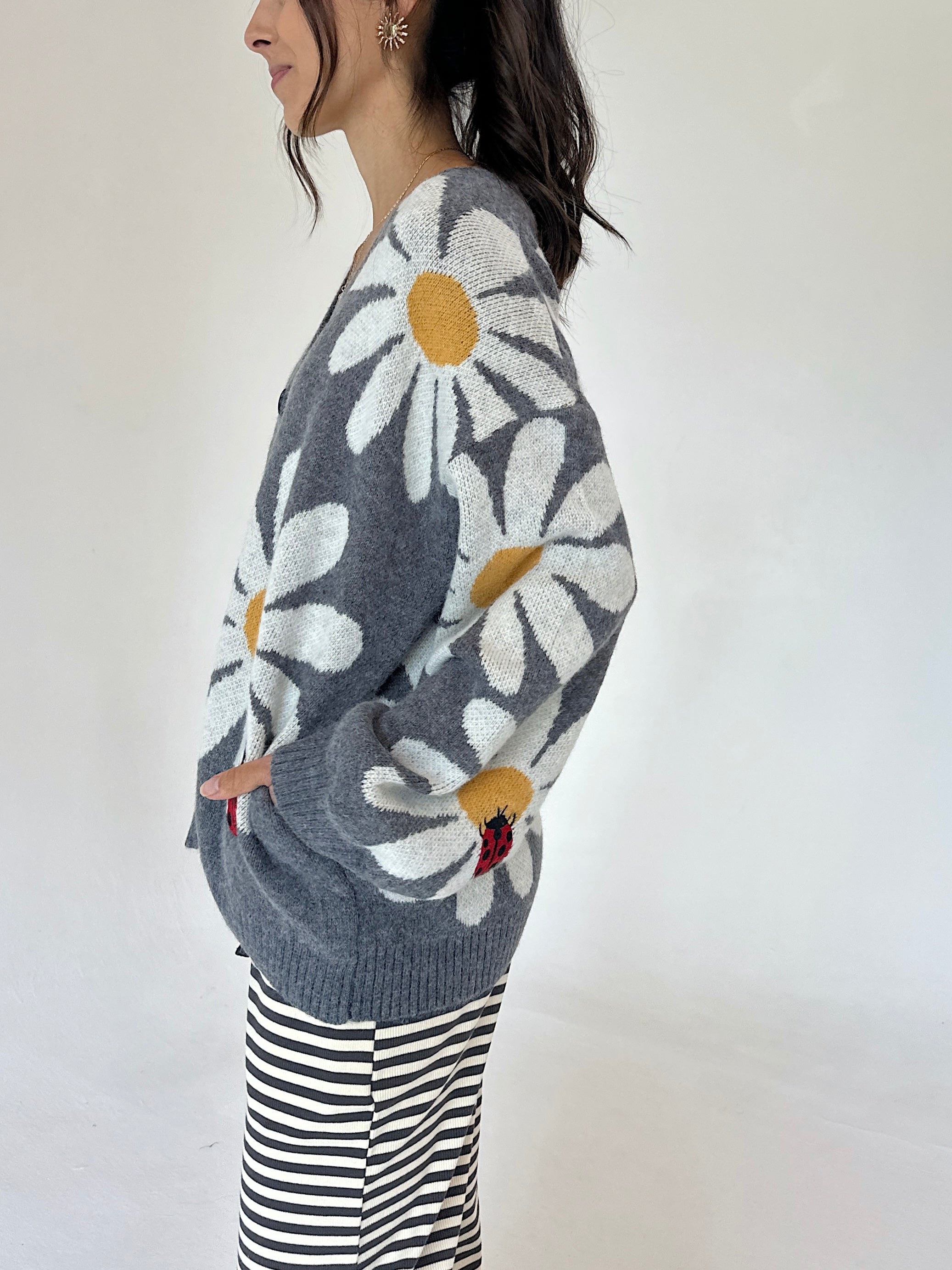 A woman wearing a gray knit cardigan with large white daisy patterns and yellow centers, paired with black and white striped pants, photographed against a plain white background.