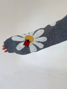 A close-up of a gray knit sleeve featuring a large white daisy with a yellow center and a red ladybug motif, held against a plain light background.