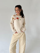 A woman modeling a cream-colored knit sweater with colorful floral embroidery on the chest, paired with yellow and white gingham wide-leg pants, against a plain light gray studio background.