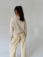 A woman is shown from behind wearing a cream-colored knit sweater and cream-and-white gingham pants against a plain light gray studio background.