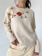 A person wearing a cream knit sweater with colorful embroidered flowers on the chest, paired with a light beige and white gingham skirt, against a plain light gray background.