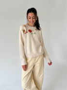 A woman modeling a cream-colored knit sweater with colorful floral embroidery on the chest, paired with cream and beige gingham pants, against a plain light gray background.