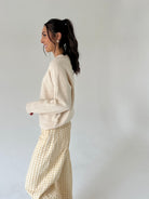 A woman in a side profile wearing a cream knit sweater and a beige and white gingham skirt, standing against a plain light gray studio background.