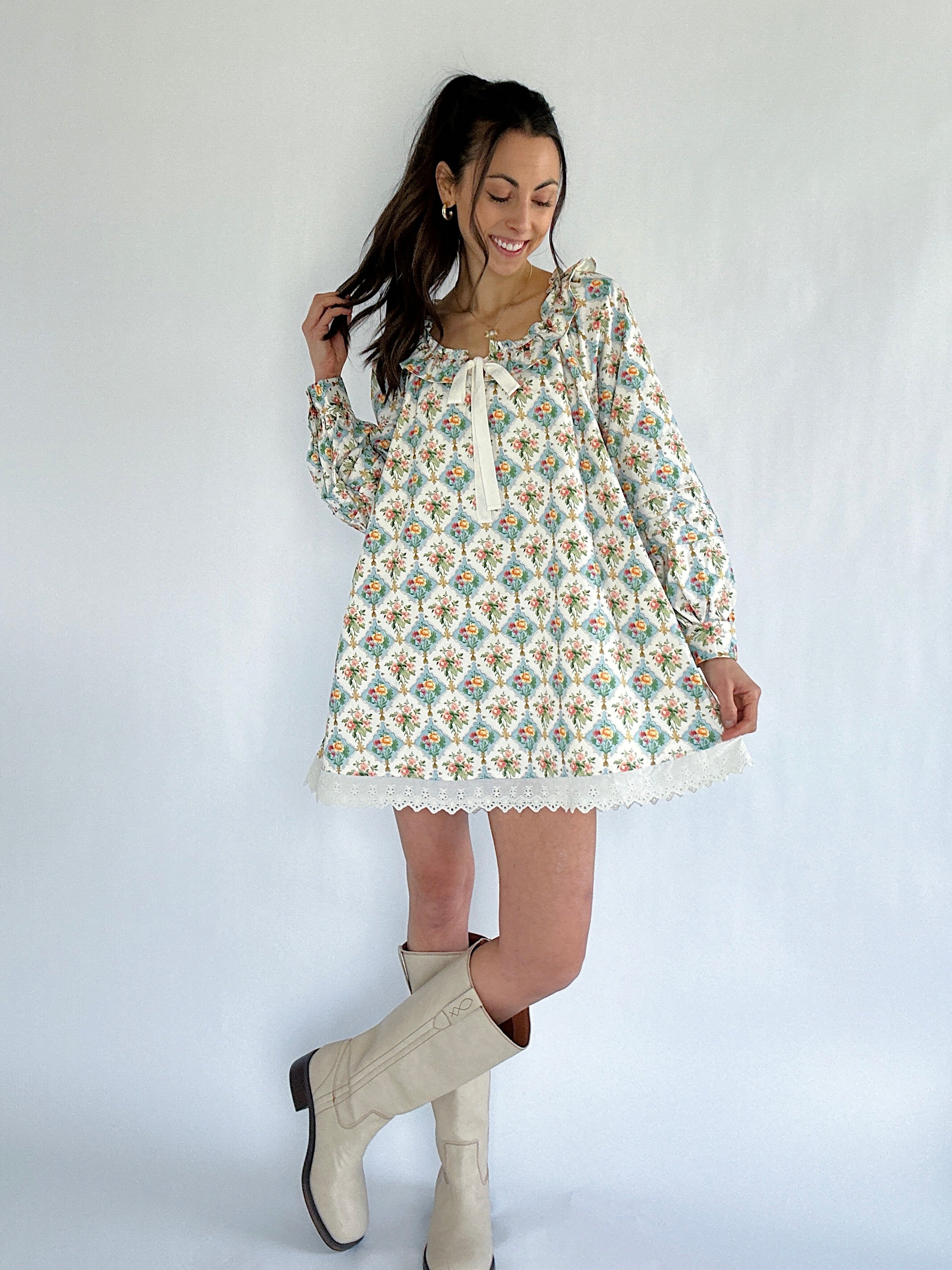 Woman wearing a floral long sleeve mini dress and white boots against a plain background