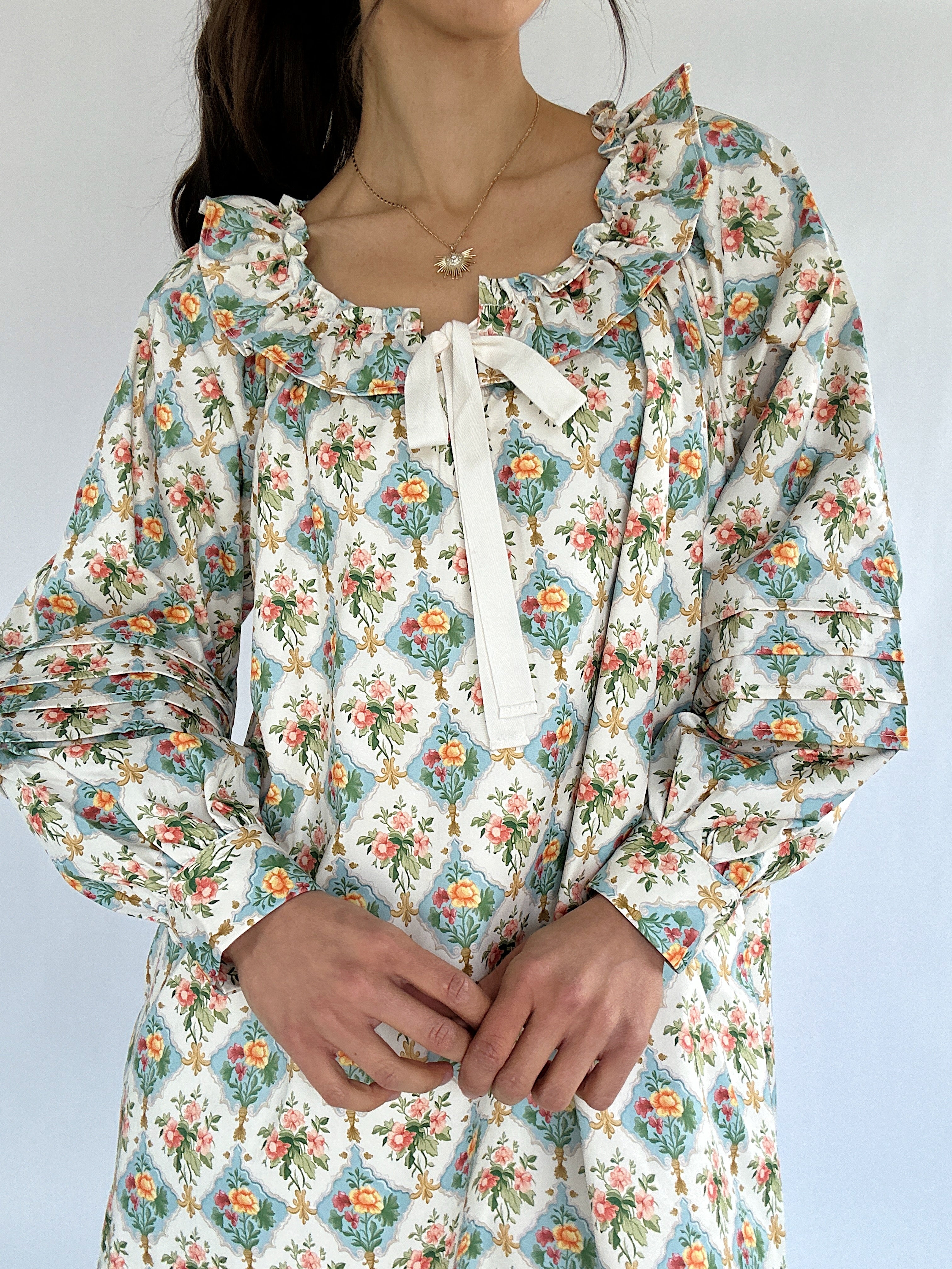 Floral patterned dress with long sleeves, a ruffled neckline and a bow tie detail on a plain background