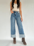 A person wearing medium washed denim jeans with a wide leg silhouette and a cuffed bottom, paired with a white sleeveless top and brown ankle boots.