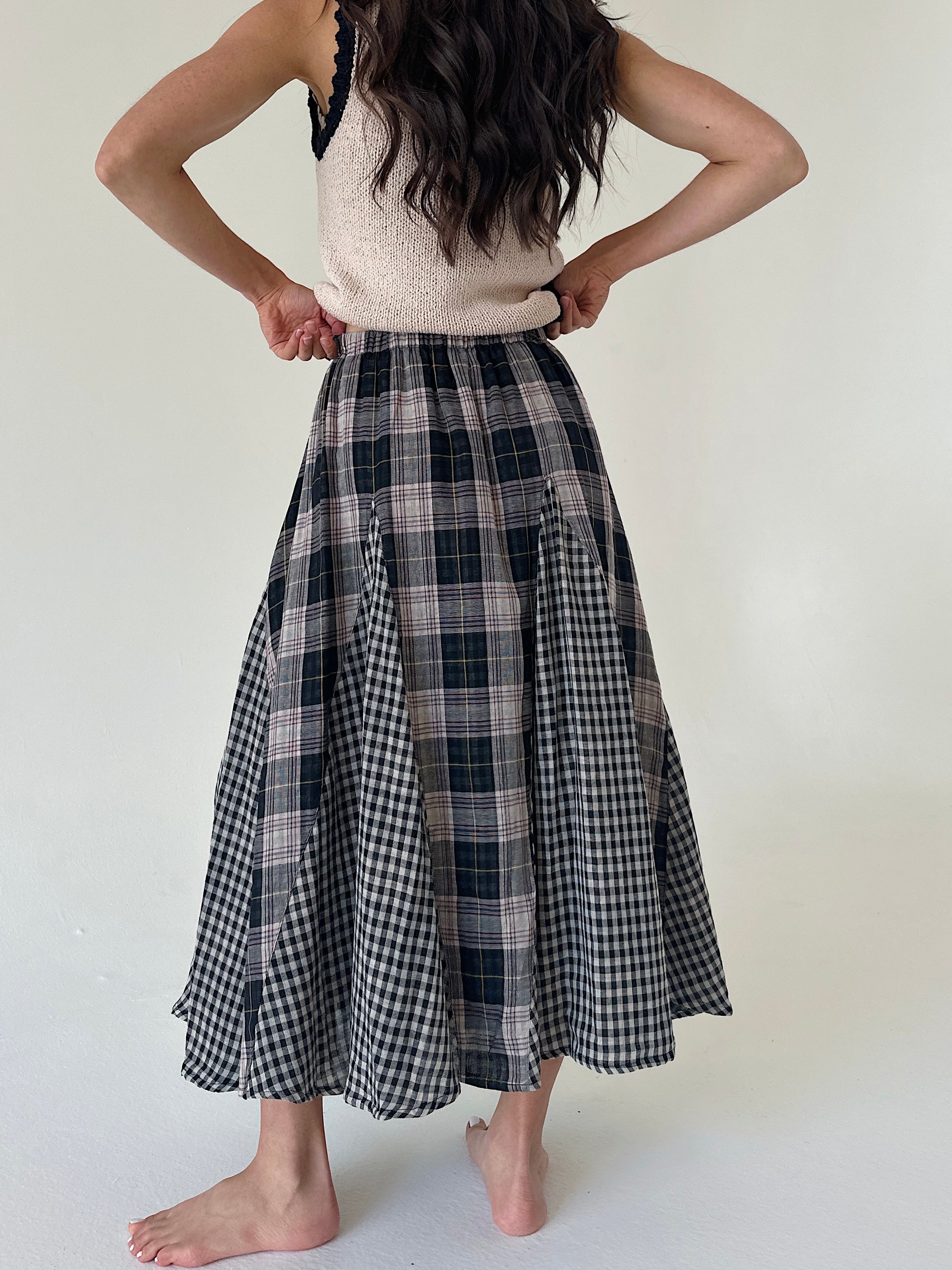 A woman shown from the back wearing a long A-line midi skirt with a mixed plaid pattern, featuring black and white gingham and dark navy/black plaid panels. She is also wearing a cream knit top, standing barefoot against a plain off-white background.