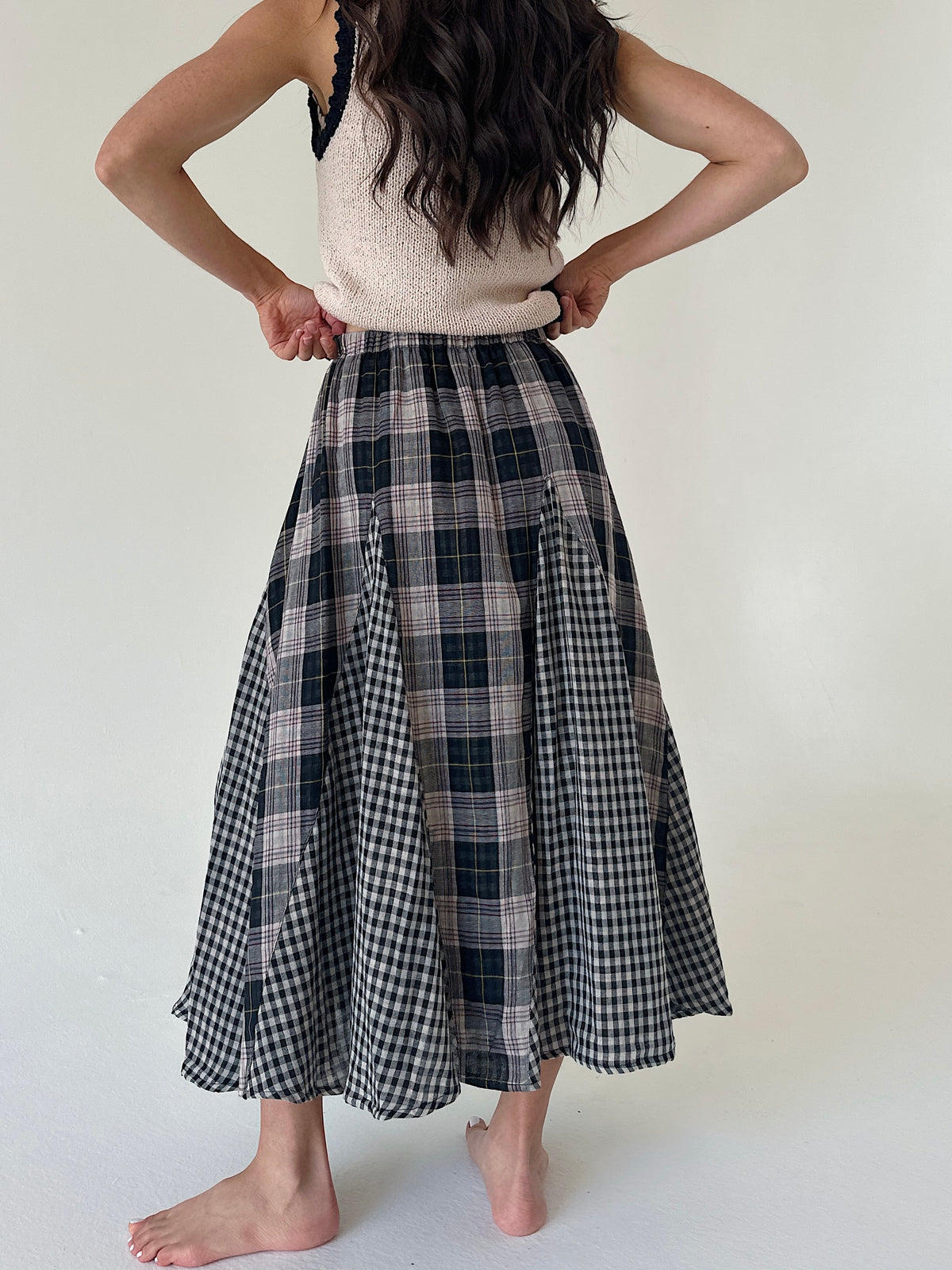 A woman shown from the back wearing a long A-line midi skirt with a mixed plaid pattern, featuring black and white gingham and dark navy/black plaid panels. She is also wearing a cream knit top, standing barefoot against a plain off-white background.