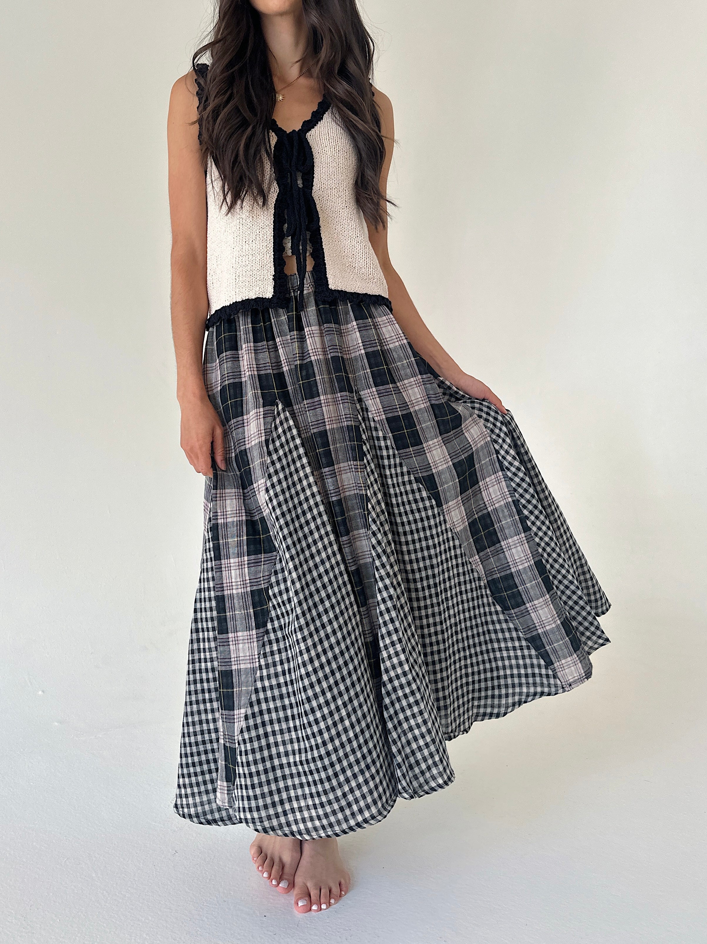 A woman modeling a sleeveless knit cream knit top and a black, gray, and white plaid skirt, standing against a plain off-white background.
