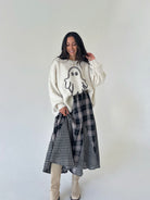 A woman stands against a plain light gray studio background wearing a cream knit sweater with a ghost graphic, a black-and-white plaid skirt, and beige knee-high boots.