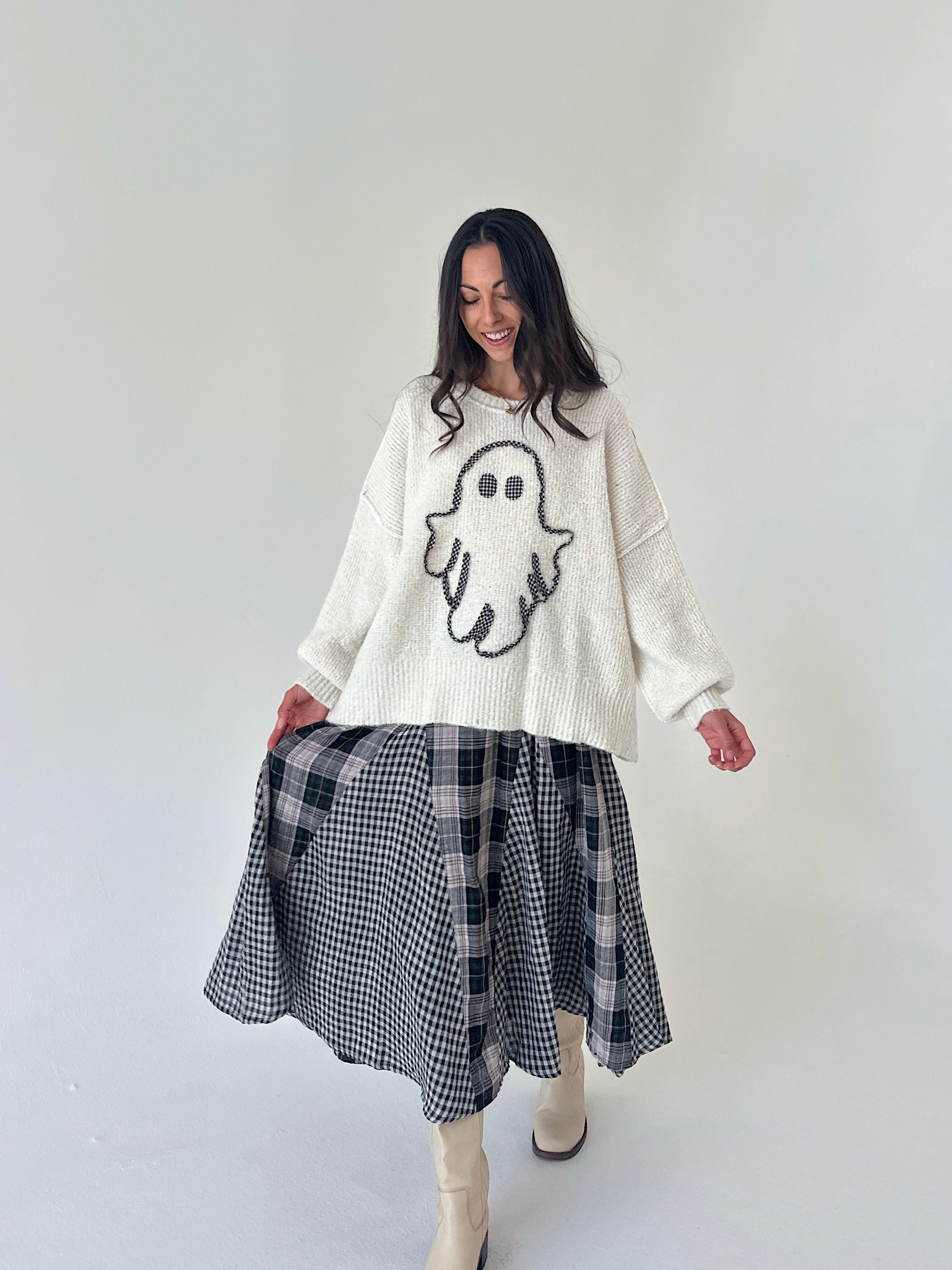 A woman modeling a cream knit sweater with a ghost graphic and a black-and-white plaid skirt, standing against a plain light gray studio background.