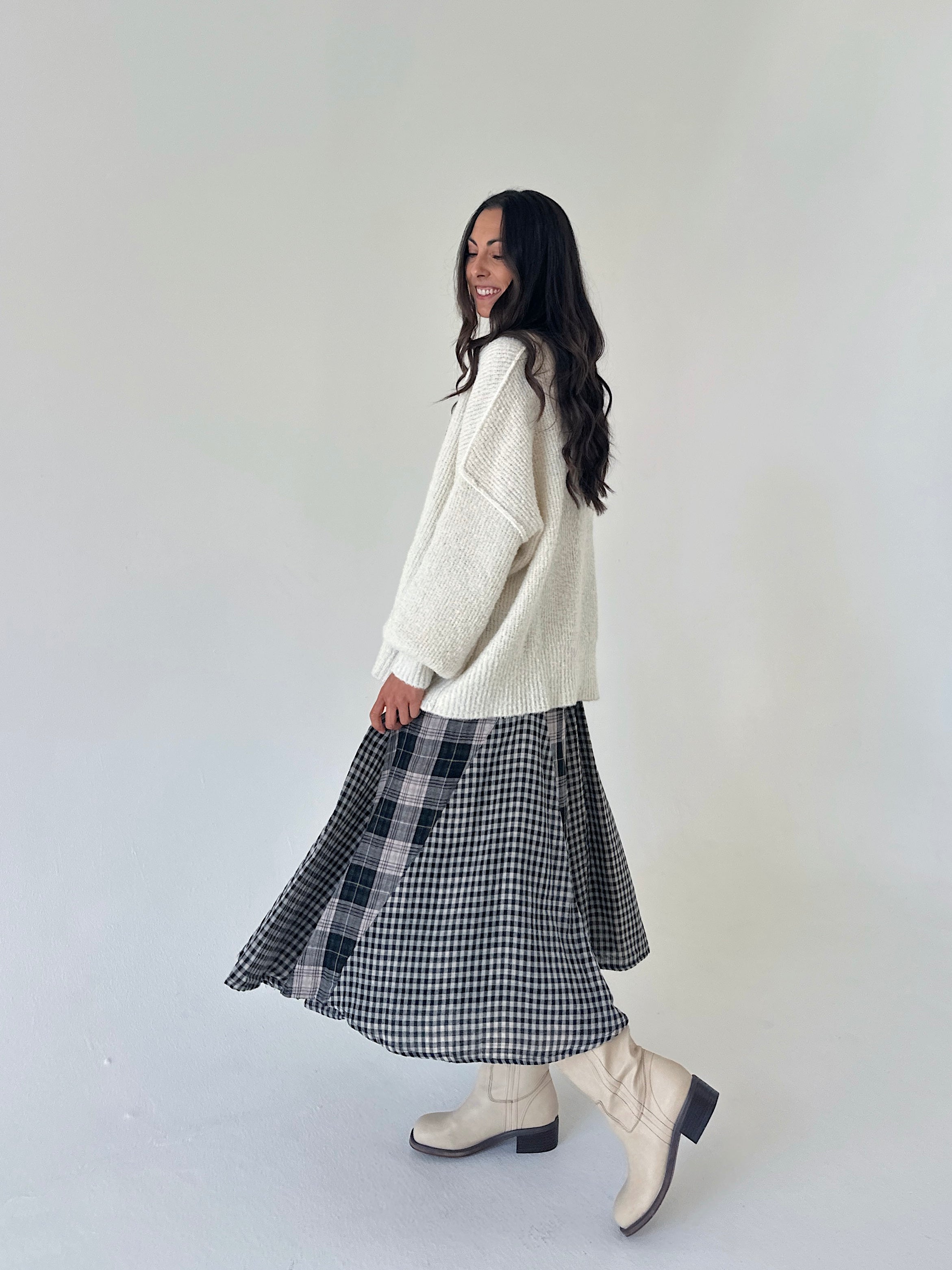 A woman wearing a cream knit sweater, a black and white gingham skirt, and cream boots, standing against a plain light gray studio background.