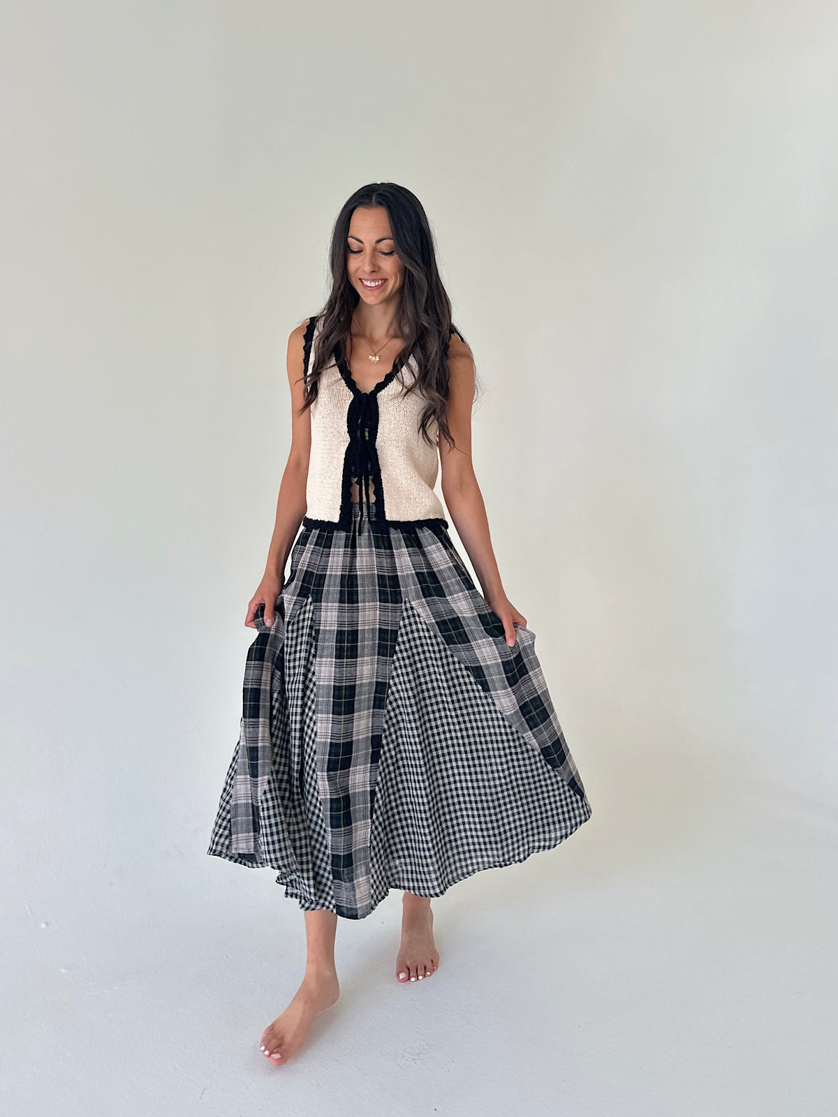 A woman modeling a sleeveless knit vest with a black and white gingham plaid skirt, standing against a plain off-white studio background.