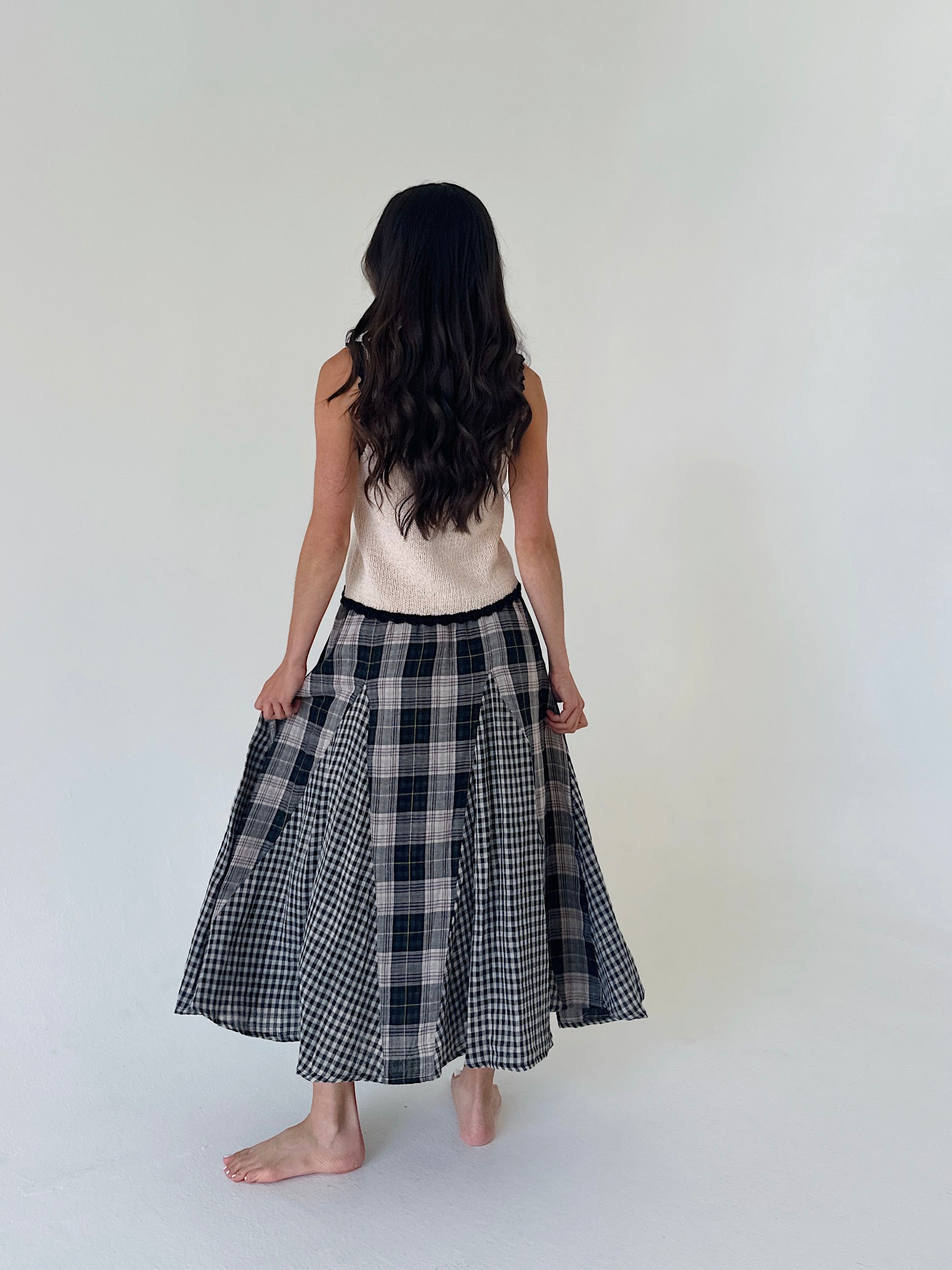 A woman viewed from behind wearing a sleeveless cream top and a black and white plaid midi skirt, standing barefoot against a plain off-white studio background.